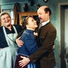 Ein Mann umarmt eine Frau, die zu ihm aufschaut. Eine ältere Frau daneben lacht fröhlich. Es wirkt häuslich und traditionell.