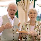 Zwei ältere Personen, die gemeinsam Würstchen servieren. Im Hintergrund eine grüne Landschaft und Menschen auf einem Fest.
