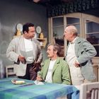 Drei Männer in Anzügen interagieren in einem Raum mit Tisch. Einer liest ein Papier, einer sitzt, der dritte schaut zu.