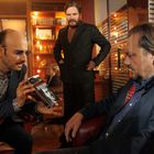 Ein Mann mit Brille zeigt ein Buch. Zwei weitere Männer sind in einem modern eingerichteten Raum.