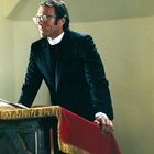 Kein Text. Ein Mann in Priesterkleidung steht hinter einem hölzernen Rednerpult mit rotem Tuch.