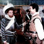 Zwei Männer in historischer Kleidung stehen sich lächelnd gegenüber, umgeben von mehreren weiteren kostümierten Personen.