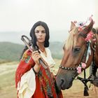 Es gibt keinen Text. Eine Frau in bunter Kleidung steht mit einem geschmückten Pferd auf einer Landschaft.