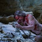 Kein sichtbarer Text. Szene: Blasser, blutüberströmter Mann mit langen Haaren in einer Höhle, in Ketten gelegt, Skelett neben ihm.