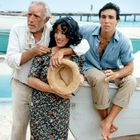 Ich kann den Text nicht erkennen. Auf einem Strand posieren drei Personen bei einem Boot. Ein Mann umarmt eine Frau, ein anderer sitzt.