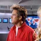 Auf einem Bildschirm im Hintergrund ist eine Weltkarte. Vordergrund: Mann mit rotem Hemd und markanten Haaren.