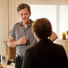 Ich kann den Text nicht lesen. Zwei Personen unterhalten sich; eine hält ein Getränk. Im Hintergrund sind Pflanzen und Gläser.