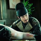 Ein Mann in traditioneller Kleidung untersucht aufmerksam einen Körper mit Wunden in einem schlichten, beleuchteten Raum.