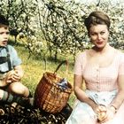 Ein Junge in Lederhosen und eine Frau in einem karierten Kleid sitzen neben einem Picknickkorb unter blühenden Bäumen.