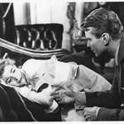 Ein Mann kniet neben einer Frau, die auf einem Sofa liegt. Er hält ihre Hand, sie schaut ihn an, während sie liegt.