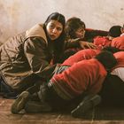 Eine Frau hockt ängstlich auf dem Boden neben Schutz suchenden Kindern in roten Pullovern an einer Wand.