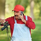 Ein Mann in rotem Hemd und Mütze, blauer Latzhose, telefoniert und hält ein Fernglas in einem Park.