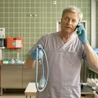 Ein älterer Mann in OP-Kleidung mit Handschuhen telefoniert und hält ein Seil in einem Krankenhauszimmer.