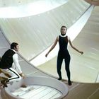 Eine Frau in einem schwarzen Anzug steht in einem futuristischen Raum. Ein Mann hockt daneben am Rand einer runden Öffnung.