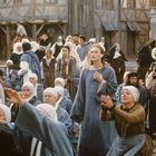Eine mittelalterliche Szene mit Menschen in einfachen Kleidern und Kopfbedeckungen auf einem Marktplatz.