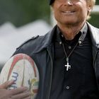 Ein Mann in Lederjacke mit Baskenmütze hält einen Rugby-Ball und trägt eine Halskette mit Kreuz.