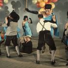 Tanzende Männer und Frauen in Tracht und blauen Kleidern, umgeben von bunten Luftballons, ein Mann fällt in eine Bodenöffnung.