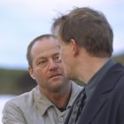 Zwei Männer im Gespräch; einer schaut ernst, der andere halb von hinten zu sehen. Sie tragen Mäntel, draußen.