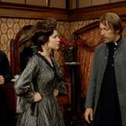 Keine erkennbaren Texte. Drei Personen in historischer Kleidung in einem viktorianisch eingerichteten Zimmer.