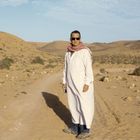 Ein Mann in weißer Kleidung und Sonnenbrille steht in einer Wüstenlandschaft mit Hügeln und vereinzelten Büschen.