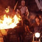 Keine sichtbaren Texte. Eine Person auf einem Motorrad fährt durch eine brennende Barriere, während eine andere Person auf einem Pferd sitzt.