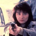 Frau mit entschlossenem Blick hält eine Pistole, trägt einen beigen Mantel. Hintergrund verschwommen und hell.