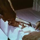 Zwei Personen bearbeiten Holz mit einer Säge. Hände halten das Material, während sie in traditioneller Kleidung gekleidet sind.