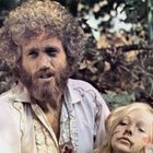 Ein Mann mit lockigem Haar und Bart sitzt im Grünen, neben ihm ein Mädchen mit blonden Haaren und Kratzern im Gesicht.