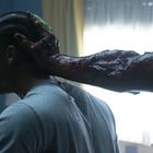 Person seitlich mit Zöpfen; eine blutverschmierte Hand greift ihren Kopf. Hintergrund unscharf, Fenster und Vorhänge sichtbar.