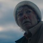 Eine ältere Frau mit besorgtem Ausdruck trägt eine weiße Mütze und einen Mantel. Der Himmel ist wolkenverhangen.