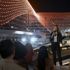 Eine Frau spricht vor Publikum mit Mikrofon. Hinter ihr ein Rennwagen mit "Vanderberg"-Aufschrift. Hintergrund von Lichtern erhellt.