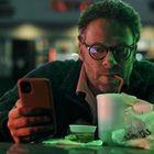 Ein Mann mit Brille schaut auf ein Handy und trinkt aus einem Becher. Vor ihm liegen Essensverpackungen.