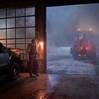 Sheriff-Fahrzeug und Mann in Garage bei geöffnetem Tor. Draußen schneit es, und ein Schneepflug steht im Nebel.
