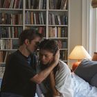 Ein Mann und eine Frau sitzen eng umschlungen auf einem Bett in einem gemütlichen Zimmer mit Bücherregal und Lampe.