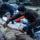 Der Text auf dem Eimer lautet: "MIEL". Zwei Personen bei einer verschneiten Flusslandschaft versorgen zwei am Boden liegende Menschen.