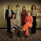 Sechs Frauen in einem Retro-Raum, zwei sitzen, vier stehen. Eine trägt einen auffälligen orangefarbenen Anzug.