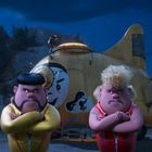 Zwei Comicfiguren in Sportanzügen stehen mit verschränkten Armen vor einem gelben Raumschiff mit aufgemalten Gesichtern.