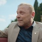 Die Person trägt einen beigen Anzug, darunter ein dunkelblaues Hemd. Er hebt begeistert die Hände. Hintergrund: Landschaft.