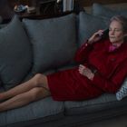 Eine ältere Frau liegt auf einem grauen Sofa, trägt einen roten Mantel und telefoniert. Mehrere Kissen umgeben sie.