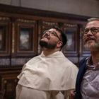 Zwei Männer stehen in einem Raum mit holzverkleideten Wänden und blicken nach oben. Einer trägt ein weißes Gewand, der andere einen blauen Anzug.