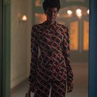 Eine Person in einem gemusterten Anzug steht in einem beleuchteten Raum, hält eine braune Tasche in der Hand.