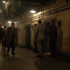 Ein düsterer Gang mit sechs Männern in Militäruniformen, die entlang alter Zellen laufen. Die Beleuchtung ist spärlich.