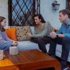 Drei Personen sitzen auf einem Sofa im Freien. Sie unterhalten sich ernsthaft; im Vordergrund ein Holztisch mit Lichthaltern.