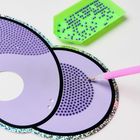 Eine lila, scheibenförmige Vorlage mit glitzerndem Rand und dunklen Punkten wird mit einem Werkzeug bearbeitet.