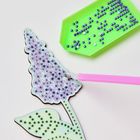 Zahlen auf einer Vorlage mit lila und grünen Schmucksteinen. Ein Stift platziert Steine auf die Blütenform.