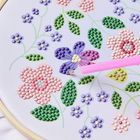 Florales Stickdesign mit rosa und lila Blumen, grünen Blättern und einem pinken Stift auf weißem Stoffrahmen.