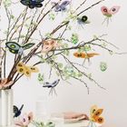 Zweige mit Filzschmetterlingen und bemalten Eiern in einer Vase, daneben gedeckter Tisch mit Gläsern und rosa Servietten.