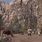 Eine dramatische Landschaft mit hohen Felsklippen, vereinzelte Bäume und eine Holzhütte, Menschen sind auf der Erde verteilt.