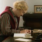 Eine Person sitzt am Schreibtisch, schreibt ins Notizbuch. Alte Fotos und Bücher sind im Hintergrund zu sehen.