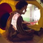 Ein Clown sitzt in einem gelben Tunnelspielplatz, gedämpft beleuchtet, mit rotem und blauem Hintergrund.